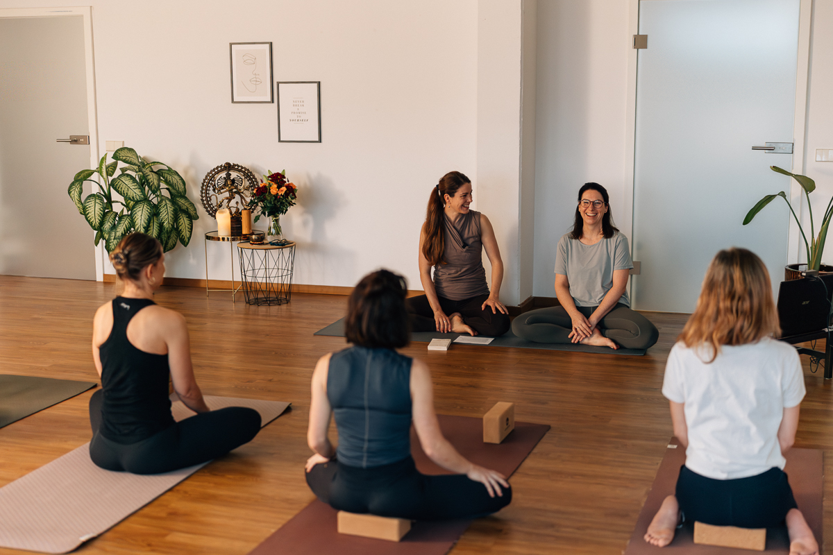 Sabine Willmann und Stefanie Altendorf sitzen gut gelaunt vor einer Yogagruppe während der Yoga Ausbildung München.